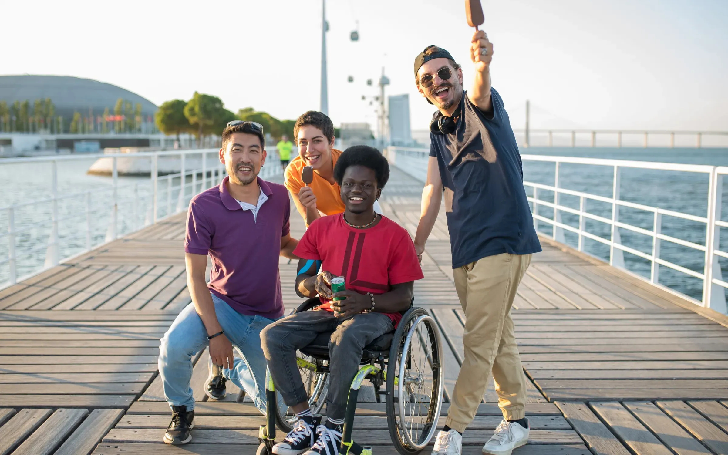 Four friends enjoying ice creams on a sunny waterfront boardwalk, one of them a wheelchair user